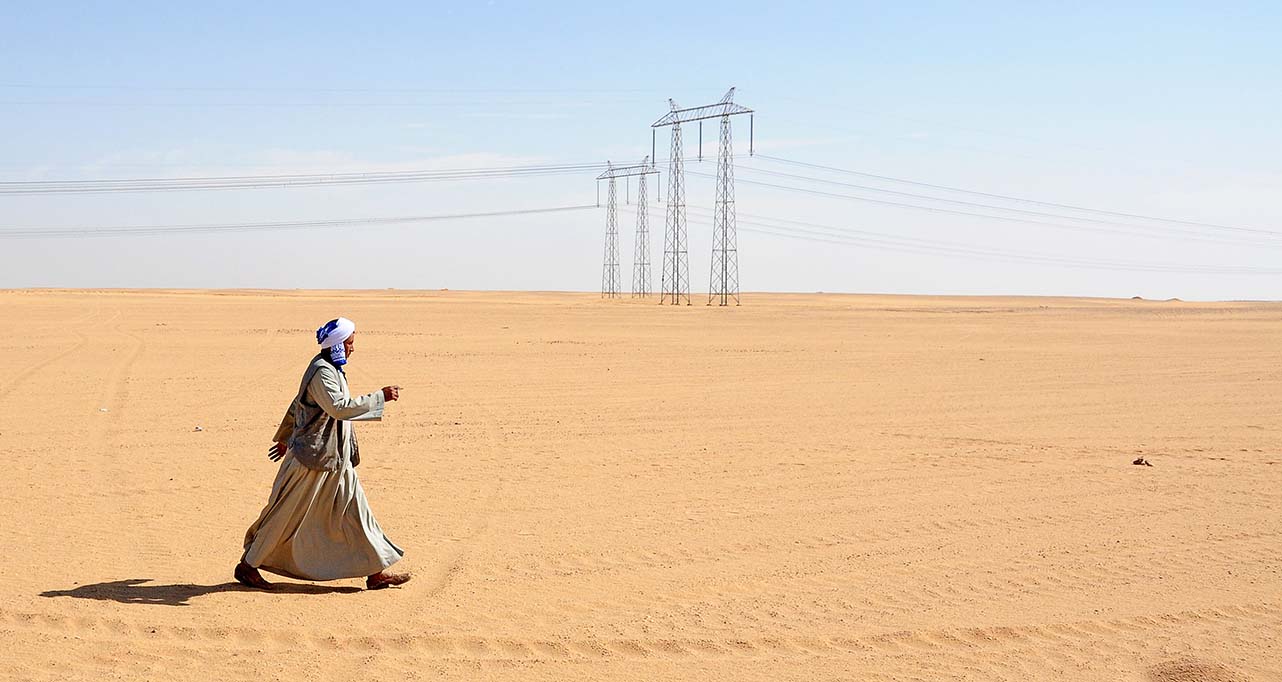 Man walking in desert next to electricity pylons